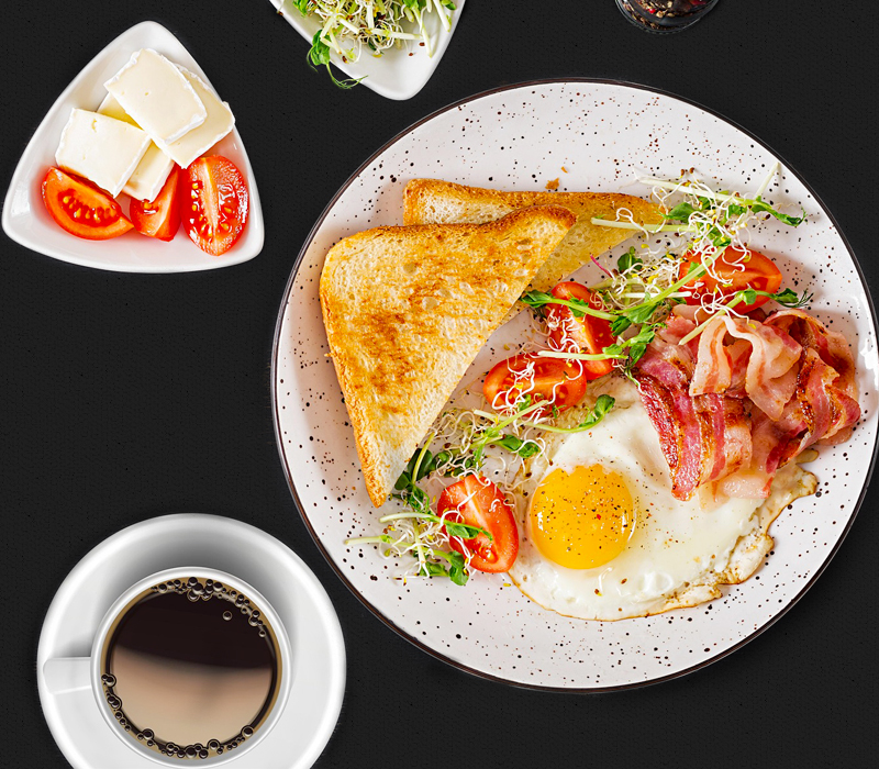 Hart an der Grenze Fruehstück Bild mit verschiedenen Frühstücksleckereien wie Ham & Eggs mit knackigem, getoastetem Brot, eine Tasse Kaffee und ein kleiner Teller mit Tomaten und Weichkäse, drapiert auf schwarzem Untergrund