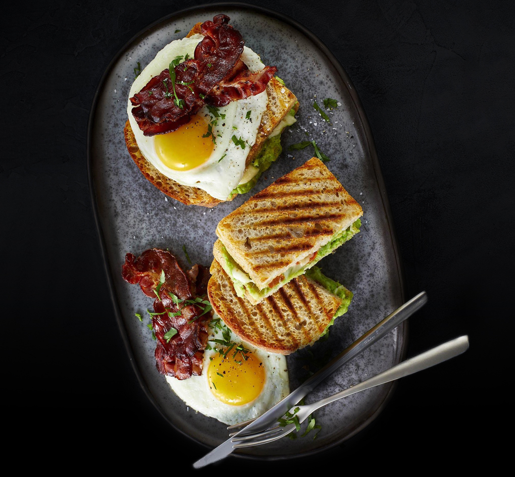 Hart an der Grenze Snacks Ovaler Teller mit einem appetitlich servierten Toast mit Spiegelei und gebratenem Speck obenauf auf dunklem Hintergrund