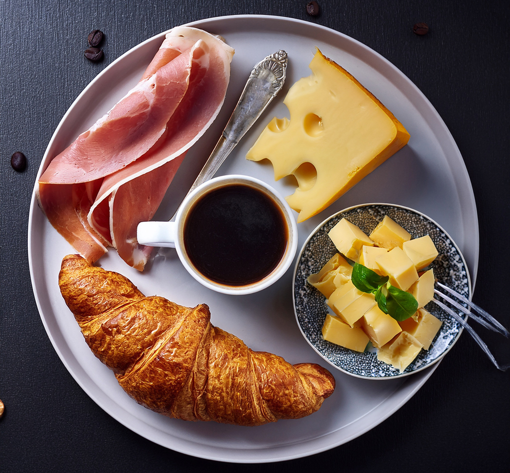 Croissant, Schinken, Käse und Kaffee appetitlich arrangiert auf einem weissen Teller mit dunklem Hintergrund