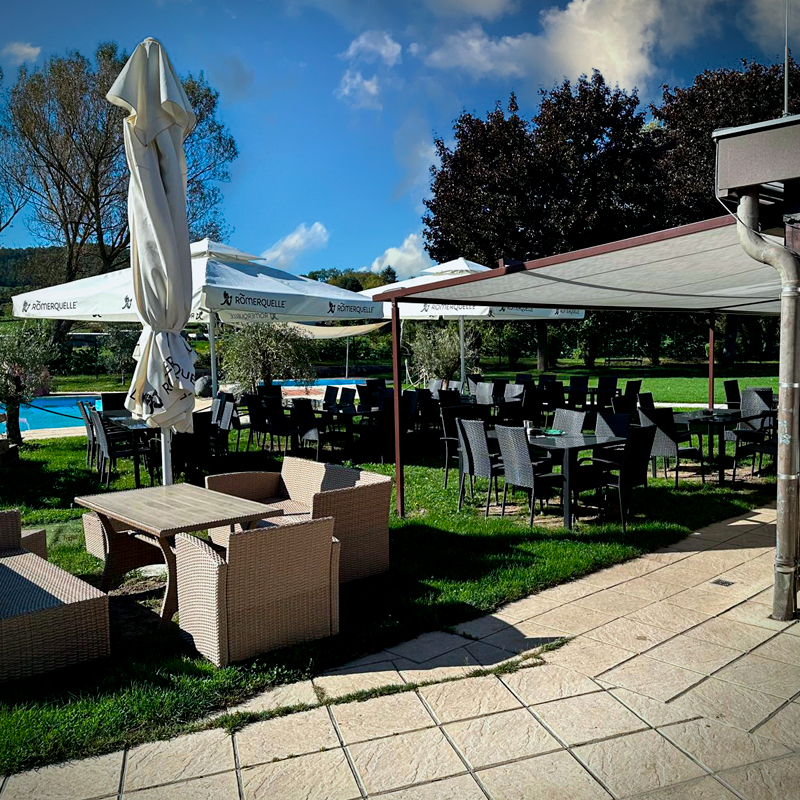 Blick auf die Terrasse mit gemütlichen Sitzgelegenheiten und Sonnenschirmen. Im Hintergrund sieht man das Freibad.